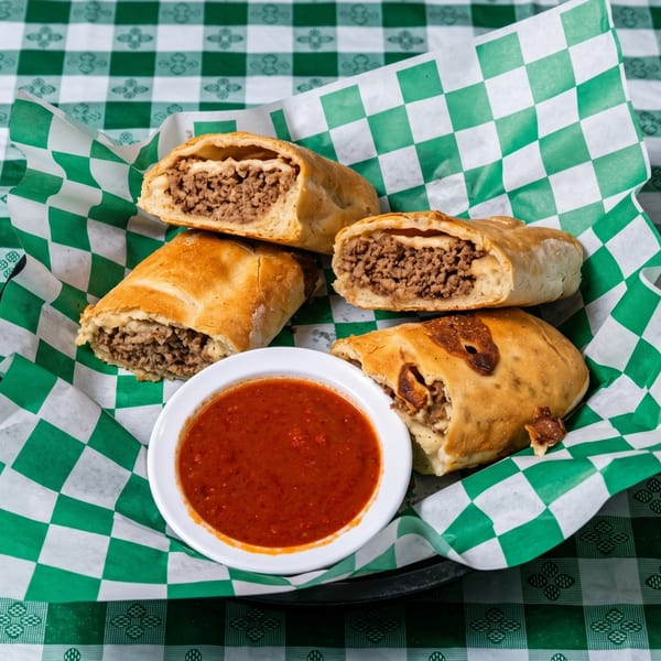 Four meat-filled calzones and a bowl of red sauce on green and white checkered paper.
