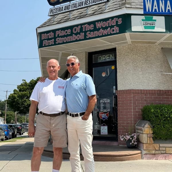 Two men stand outside a restaurant with a sign proclaiming it the "Birth Place Of The World's First Stromboli Sandwich."