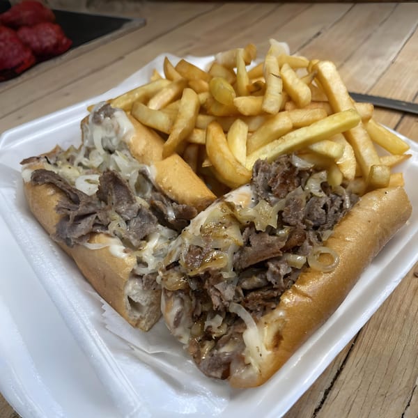 A Romano's Pizzeria cheesesteak sandwich with onions and fries in a white takeout container.