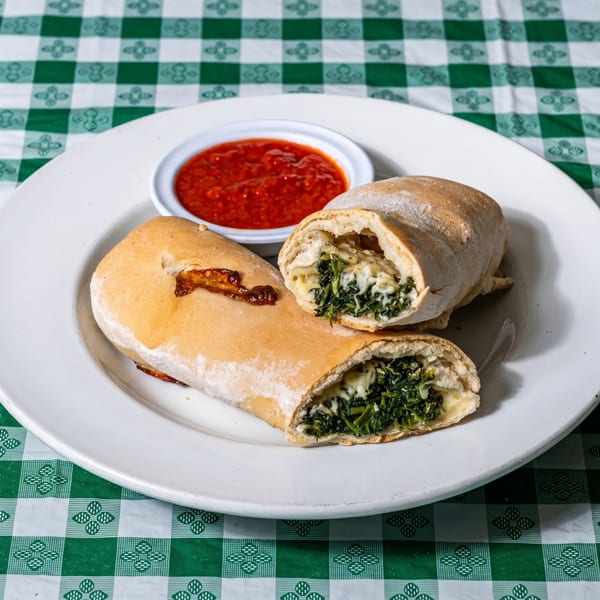 Two spinach and cheese strombolis, one cut open, with a side of marinara sauce on a white plate.