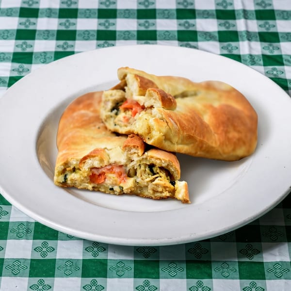 Two golden-brown calzones, one broken open to reveal chicken, tomatoes, and olives, on a white plate.