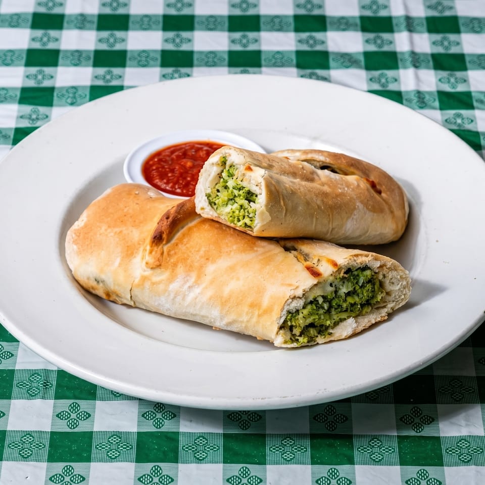 Two calzones filled with broccoli and cheese, served with a side of marinara sauce on a white plate.